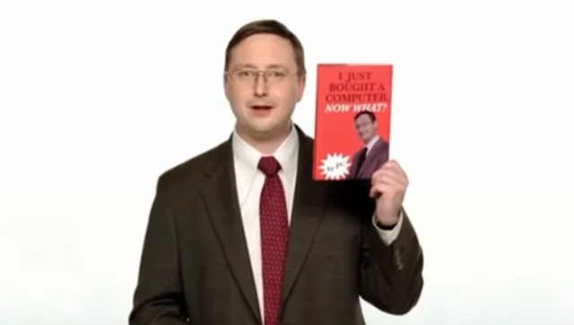 A man in a suit holding up a red book titled “I Just Bought a Computer, Now What?”